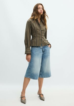 Jeune femme portant une chemise marron rayée boutonnée, un short en jean délavé au niveau des genoux et des talons gris pointus, debout avec une main dans la poche.