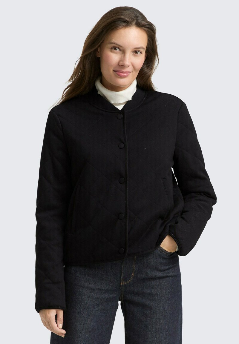 Czarna pikowana kurtka z okrągłym dekoltem, z przodu zapinana na guziki, z bocznymi kieszeniami, noszona na białym golfie. Wykonana z gładkiej, teksturowanej tkaniny.