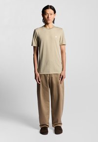 Ein leicht olivgrünes gemustertes T-Shirt mit Rundhalsausschnitt, kombiniert mit lockeren hellbraunen Hosen und braunen Slippern, getragen von einem stehenden männlichen Modell.