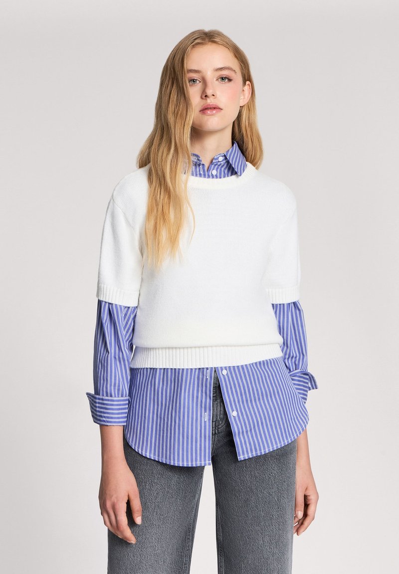 Biały krótki sweter z dzianiny z krótkim rękawem nałożony na niebieską w biały pasek koszulę z guzikami, w połączeniu z ciemnoszarymi dżinsami. Sweter ma żebrowany dół.