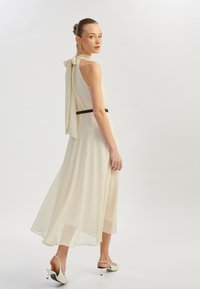 Vaaleanbeige mekko, jossa on pilkkuja, halterneck-olkaimet solmittuna laatikoksi, virtaava A-linjainen hame ja musta vyö. Valkoiset slingback-korkokengät viimeistelevät asun.