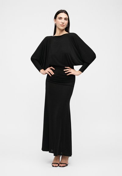 Γυναίκα που στέκεται με τα χέρια στη μέση, φοράει μακριφό μαύρο φόρεμα με μανίκια νυχτερίδας και μαύρα πέδιλα με ανοιχτά δάχτυλα, δίπλα σε απλό λευκό φόντο.