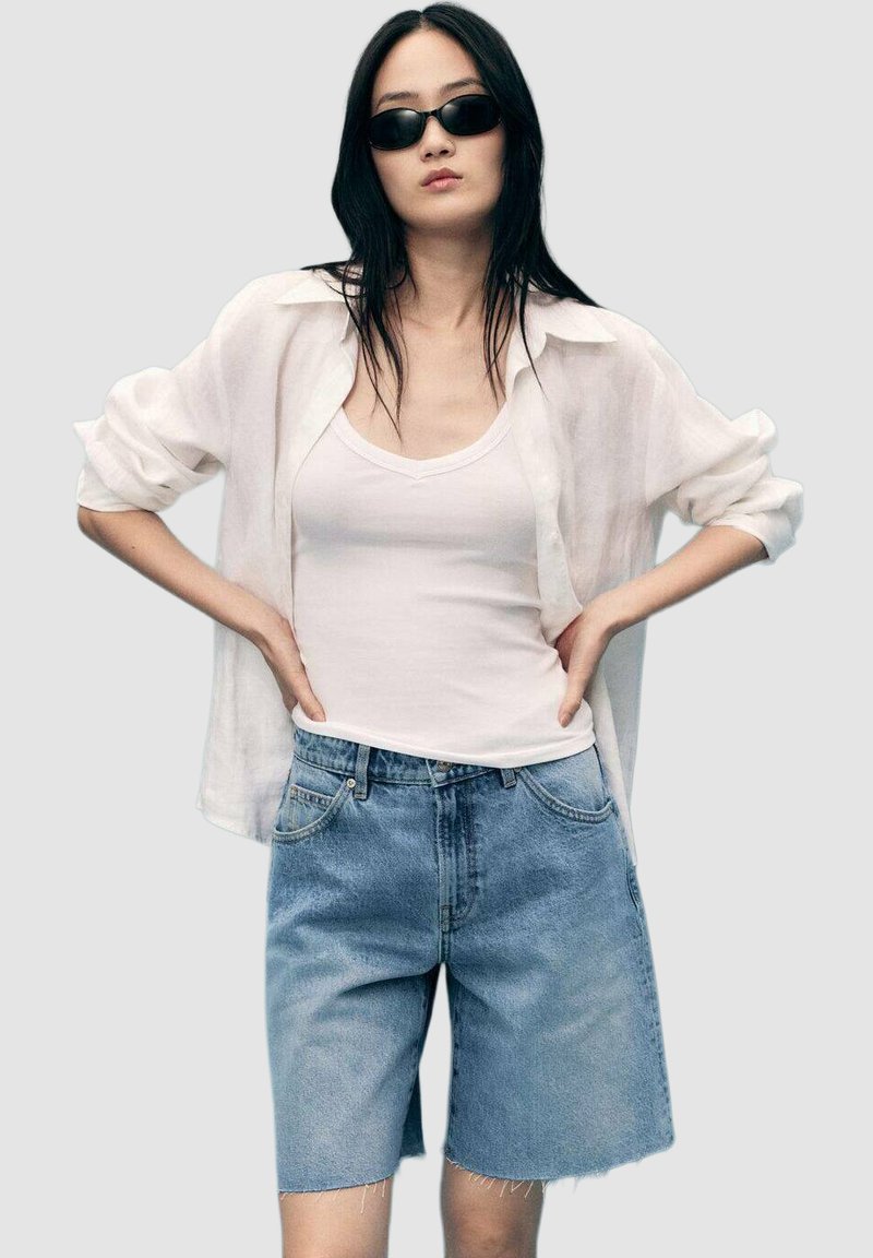 Biely long-sleeve tričko s bielym topom s tenkými ramienkami je vrstvené spolu s modrými džínsovými šortkami. Oblečenie má voľný strih a mierne roztrhané okraje.