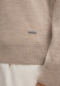 Beige gestrickter Pullover mit gerippten Bündchen und Saum, der ein metallisches Logoschild trägt. Weiche Textur mit glatter Oberfläche.