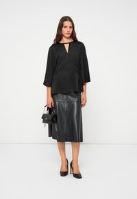 Vrouw in een zwarte top met een sleutelgatdecolleté, een zwarte leren midi-jurk, zwarte hakken en met een kleine zwarte handtas met gouden ketting.