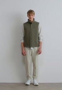 Oliwkowa zielona kamizelka bez rękawów na białej koszuli, w połączeniu z jasnobeżowymi spodniami i białymi tenisówkami; minimalistyczny design, subtelna tekstura.