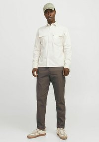 Helles beige Hemd mit Knopfleiste und zwei Brusttaschen, kombiniert mit dunkelbraunen Hosen und beigen Sneakers, ergänzt durch eine hellgrüne Kappe.