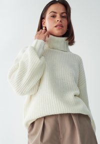 Maglione spesso bianco lavorato a maglia con colletto a coste alto, vestibilità rilassata e spalle scese, abbinato a pantaloni plissettati marrone chiaro.