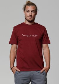 T-shirt en coton bordeaux avec un design graphique blanc au texte "So einfach ist das" sur la poitrine, manches courtes et col rond.
