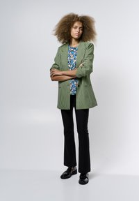 Blazer a scacchi verde e nero con rever a lancia sopra una camicia blu floreale, abbinato a pantaloni a campana neri e mocassini neri.