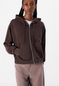 Felpa marrone con zip, cappuccio regolabile e tasche frontali, con polsini a costine. Realizzata in materiale morbido, abbinata a pantaloni marroni chiari.