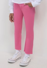 Person, der leuchtend pinkfarbene Hosen mit rohen Säumen, weiße Socken, weiße Turnschuhe und ein weißes Langarmoberteil trägt, steht vor einem hellen Hintergrund.