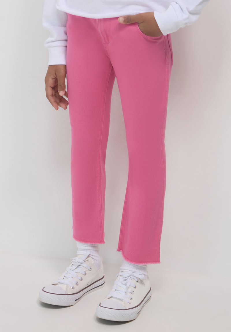 Person, der leuchtend pinkfarbene Hosen mit rohen Säumen, weiße Socken, weiße Turnschuhe und ein weißes Langarmoberteil trägt, steht vor einem hellen Hintergrund.