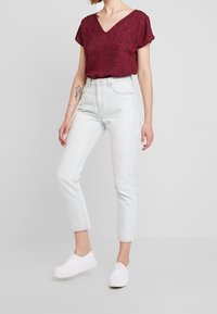 Kvinde iført en bordeauxmønstret kortærmet bluse, højtaljede lyseblå jeans, hvide plateau sneakers og blomster-tatoveringer på sin arm.
