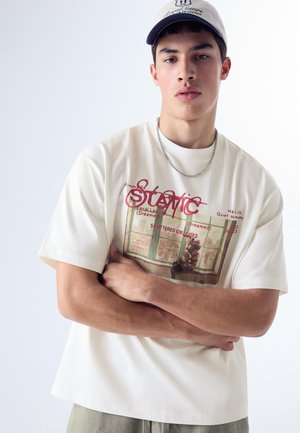 Mladý muž, ktorý má na sebe biele tričko s grafikou a nápisom „Static“, reťazový náhrdelník a svetlú čiapku na jednoduchom pozadí.