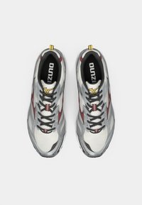 Gray and white athletic shoes with a mesh upper, black laces, red accents, and a yellow pull tab on the heel. Features a textured sole.