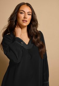Blusa nera con scollatura a V caratterizzata da dettagli in cucitura bianca. Maniche lunghe e gonfie e tessuto morbido e fluido. Texture liscia.