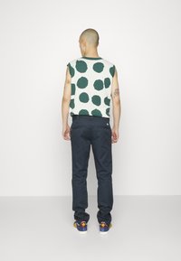 Maglione senza maniche con pois verdi su sfondo bianco, abbinato a pantaloni blu scuro e sneakers multicolori.