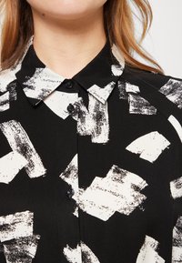 Czarna koszula z białym abstrakcyjnym wzorem pędzla, z kołnierzykiem i zapięciem na guziki z przodu. Gładka tekstura materiału.