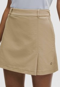 Jupe beige longueur genou avec pli sur le devant, passants pour ceinture, poches latérales et petit logo rond près de l'ourlet portée avec un haut blanc.