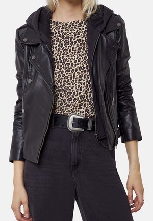 Kobieta w czarnej skórzanej kurtce nałożonej na czarną bluzę z kapturem, bluzce w panterkę, czarnych dżinsach i czarnym pasku z srebrną klamrą.