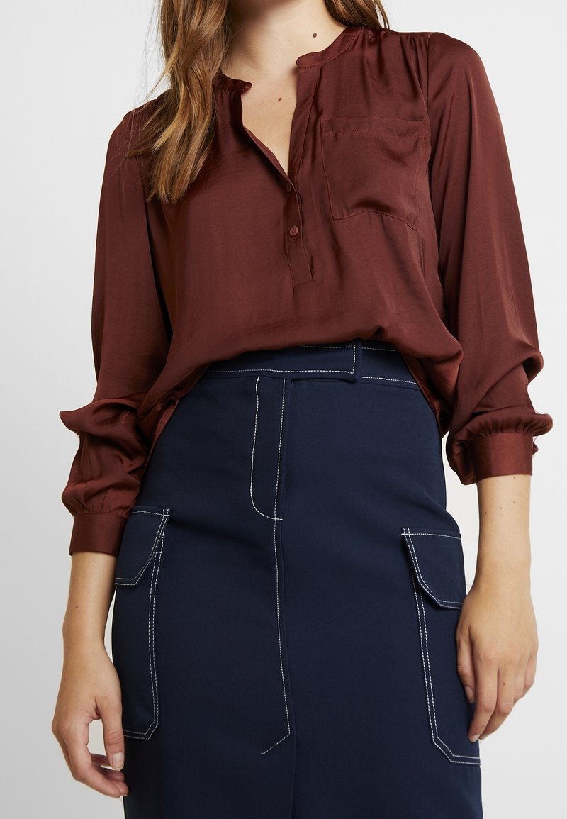 Femme portant un chemisier bordeaux boutonné rentré dans une jupe bleu marine avec de grandes poches avant et des détails de surpiqûres blanches.