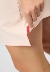 Lichtroze shirt tentoongesteld, met een textuurstof en een klein rood labelaccent aan de zijnaad, gedeeltelijk van de hand getrokken.