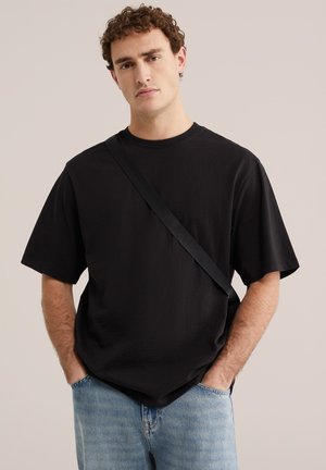 Junger Mann mit lockigem Haar, trägt ein schlichtes schwarz übergroßes T-Shirt, hellblaue Jeans und einen schwarzen Umhängegurt, die Hände in den Taschen.