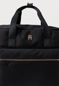 Mochila preta em nylon com duplas alças superiores, apresentando um fecho de correr dourado e um emblema central. Textura suave com um design elegante.