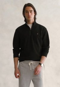 Jongeman staat in een zwart zip-up sweater, witte onderhemd, grijze joggingbroek met trekkoord en een horloge tegen een effen achtergrond.