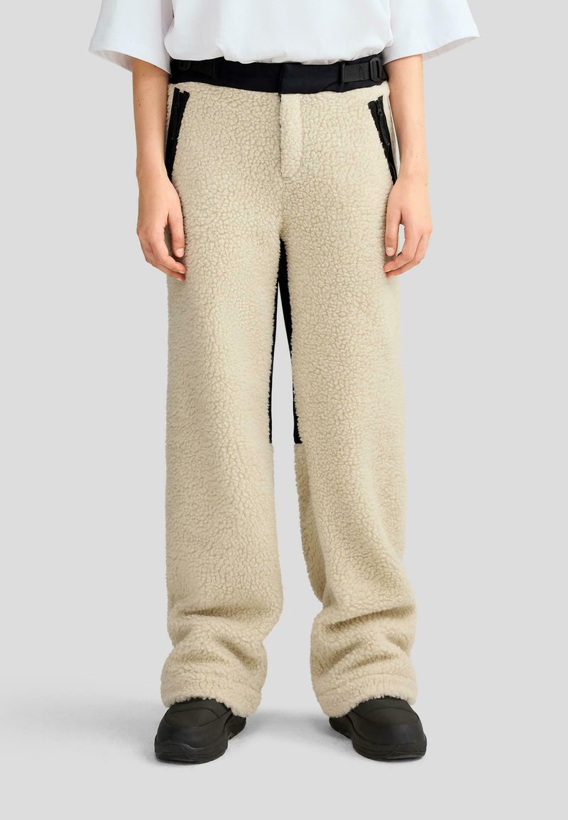 Pantalones de forro polar beige con un acabado texturizado, que presentan paneles laterales negros y un cinturón ajustable negro. Corte holgado con puños en los bajos.