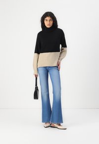 Pull à col roulé noir et beige avec une texture côtelée, associé à un jean clair évasé bleu et des chaussures beige à enfiler. Sac à main noir inclus.
