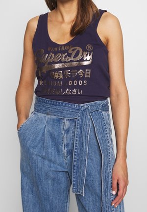 Femme portant un débardeur bleu marine avec le texte doré « SuperDry » et un pantalon en jean bleu taille haute avec ceinture nouée, les mains dans les poches.