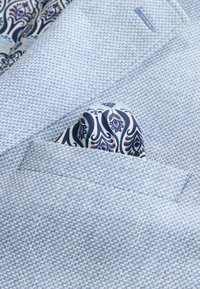 Lichtblauw gebreid blazerzakje met een gedeeltelijk zichtbaar zakdoekje in marineblauw, wit en oranje met een paisley-patroon.