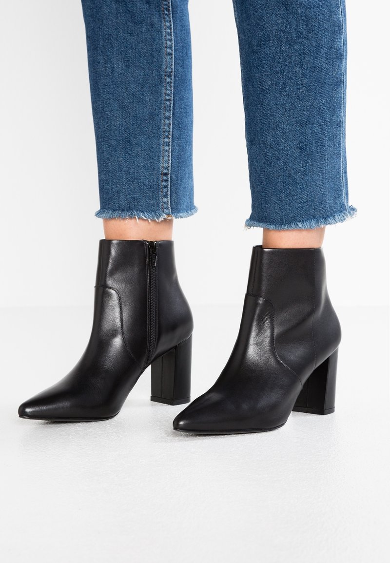 Bottes noires en cuir à talons carrés portées avec un jean bleu raccourci aux ourlets effilochés sur fond blanc.