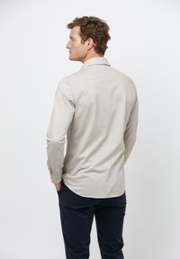 Langemouwen, lichtbeige shirt met een puntenkraag, met een effen textuur en knoopmanchetten, gedragen met donkerblauwe broek, getoond van achteren.