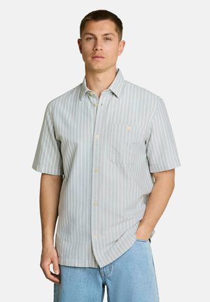 Homme portant une chemise à manches courtes boutonnée à rayures verticales gris clair et blanc avec une poche poitrine, et un jean bleu clair.