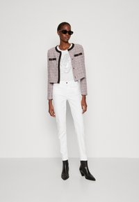 Giacca in tweed rosa e grigia con dettagli neri, bottoni dorati, abbinata a una t-shirt bianca con grafica e pantaloni bianchi slim-fit, e stivaletti neri alla caviglia.
