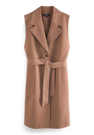 Persona con abrigo marrón sin mangas con cinturón sobre top y pantalones negros, de pie en una calle de la ciudad cerca de un coche negro y edificios.