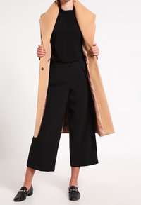 Beige coat with wide lapels and shiny interior, paired with a black blouse and wide-leg black trousers, complemented by black loafers.