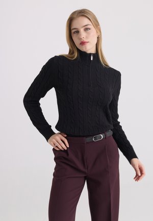 Femme aux cheveux blonds raides portant un pull en maille torsadée noir avec fermeture éclair et un pantalon violet foncé avec ceinture noire, debout devant un fond uni.