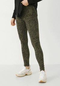Leggings vert foncé avec un motif à pois dans des teintes plus claires, fabriqués en tissu extensible et assortis à des baskets blanches.