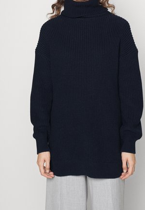 Pull à col roulé en maille épaisse bleu marine avec manches longues porté avec un pantalon gris clair sur une personne debout devant un fond neutre.