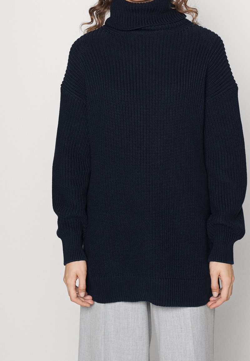 Pull à col roulé en maille épaisse bleu marine avec manches longues porté avec un pantalon gris clair sur une personne debout devant un fond neutre.