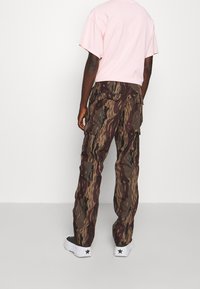 Spodnie cargo w kamuflażu w odcieniach brązu i oliwkowego, z wieloma kieszeniami, zestawione z jasnoróżowym t-shirtem i białymi trampkami.