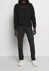 Svart huvtröja med ficka framtill, med rosa text, kombinerad med mörka jeans och vita sneakers. Slät tyg, lös passform.