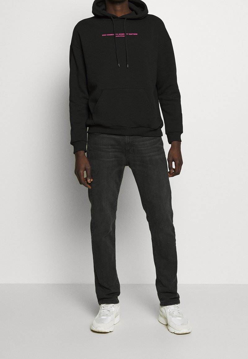 Svart huvtröja med ficka framtill, med rosa text, kombinerad med mörka jeans och vita sneakers. Slät tyg, lös passform.