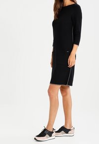 Abito di maglia nero con maniche a tre quarti e due tasche frontali, abbinato a scarpe da ginnastica nere e rosa con materiali a contrasto.