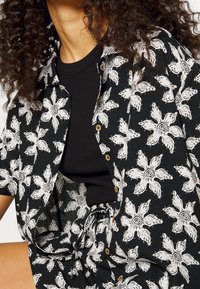 Osoba w czarnej koszuli pod czarną koszulą zapinaną na guziki oraz szortach z dużymi białymi spiralnymi kwiatowymi wzorami, z widocznymi na brzegach kręconymi włosami.