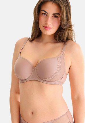 Femme aux longs cheveux bruns portant un soutien-gorge mauve avec un détail en dentelle, debout devant un fond clair uni.